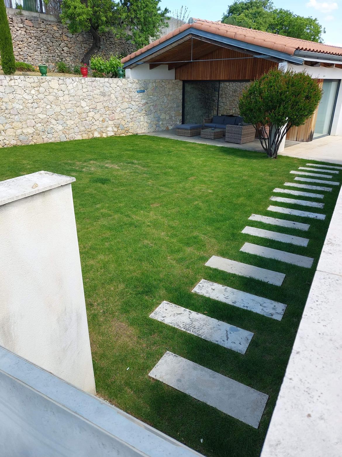 Maison contemporaine avec jardin vert luxuriant, allée de dalles en pierre et terrasse aménagée, adossée à un mur en pierre.