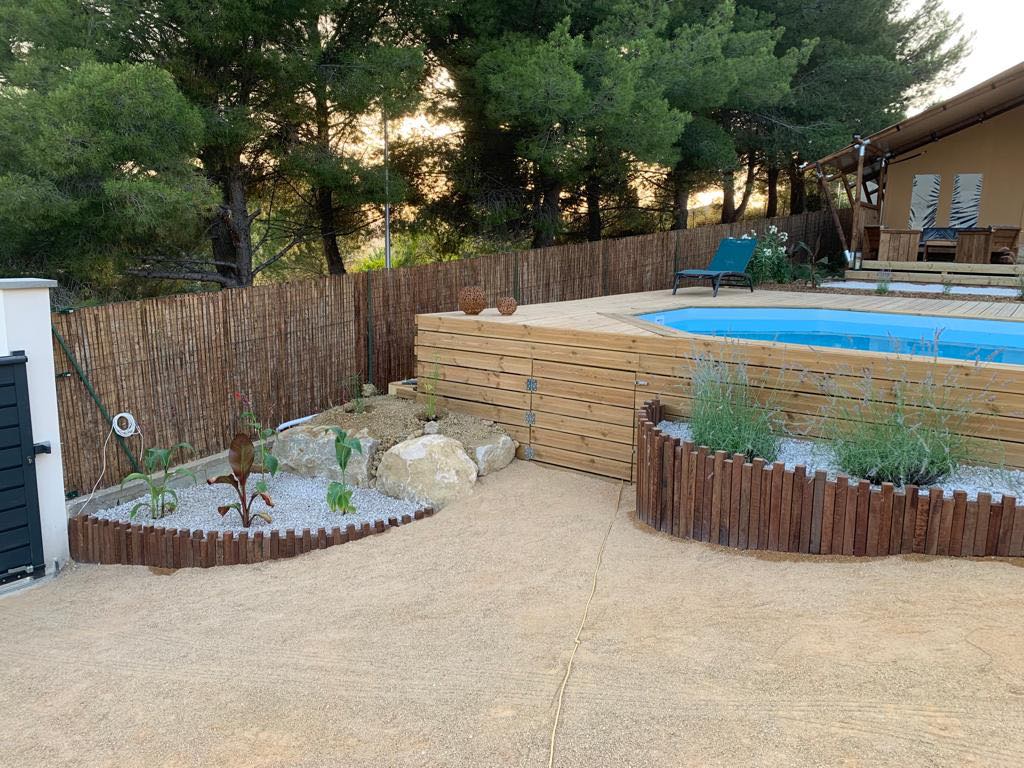 Espace extérieur avec piscine et terrasse en bois, jardin paysager, clôture bambou et tente glamping au coucher du soleil.