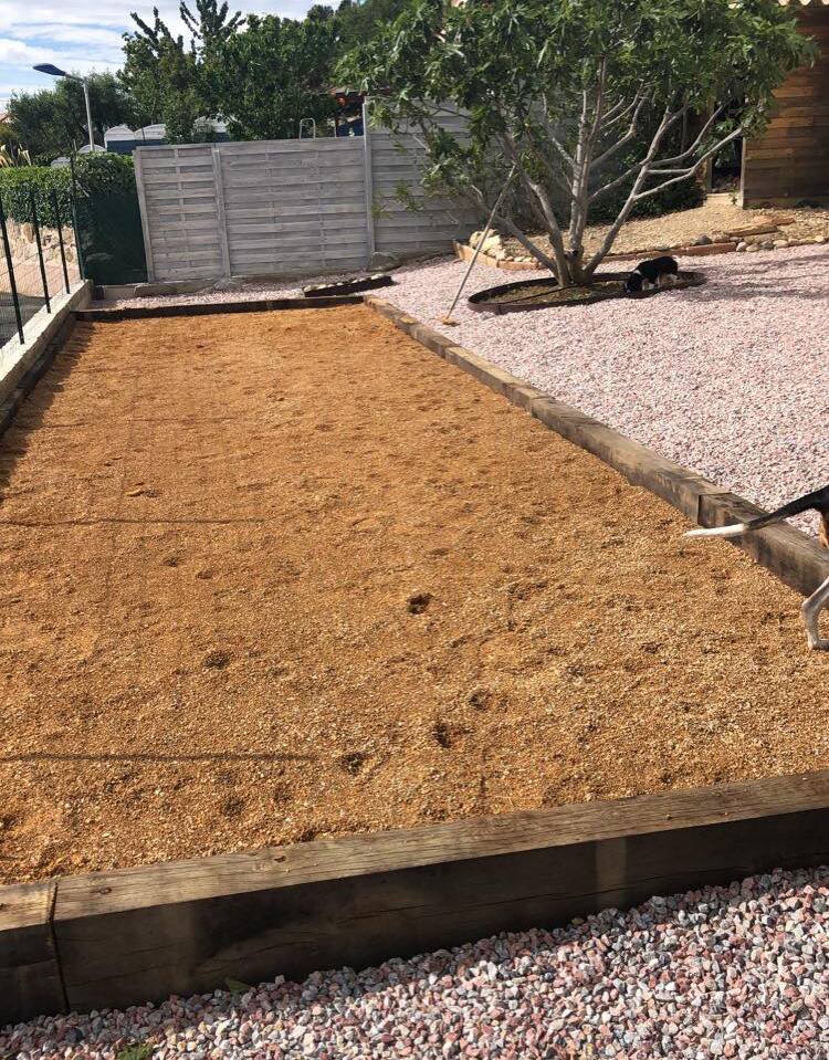 Terrain de pétanque en sable bordé de bois dans un jardin paysager avec gravier clair, arbre et petit chien noir.