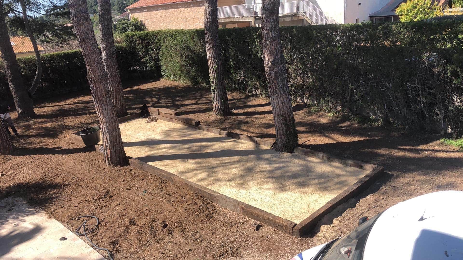 Construction d'une aire de pétanque en bois et gravier clair entourée de pins et d'une haie dense ensoleillée.