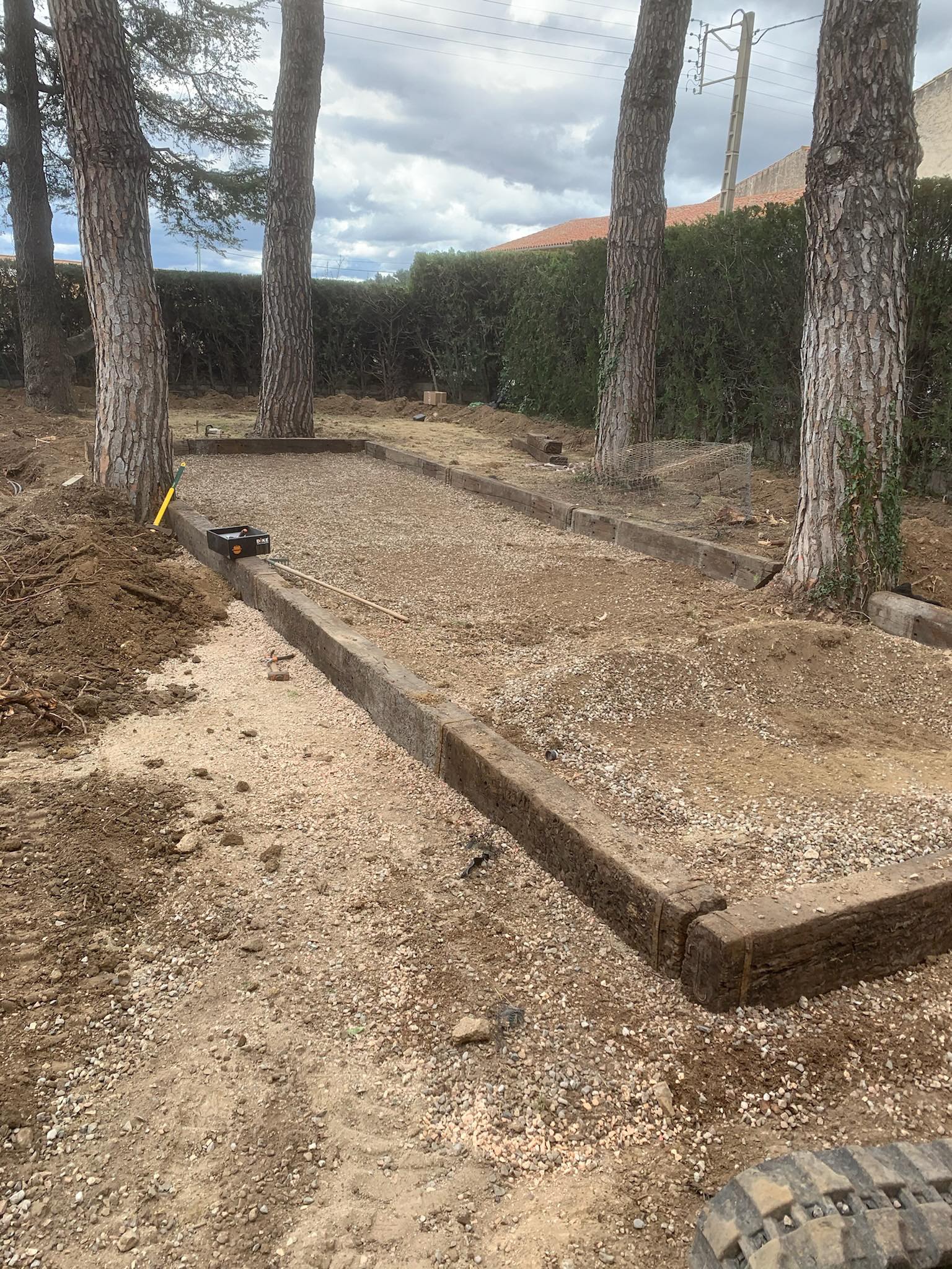 Construction d'un terrain de pétanque avec traverses en bois, gravier, entouré de pins et d'une haie, sous un ciel nuageux. Boîte d'outils noire avec texte 'DIKE' visible.